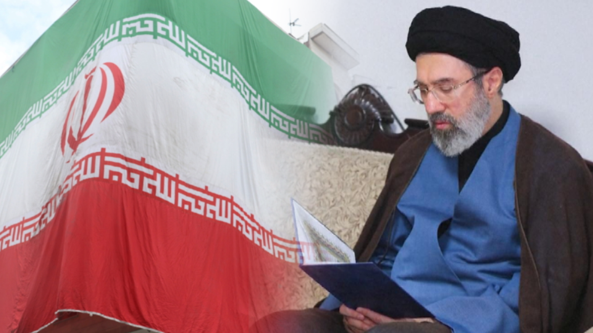 Mężczyzna w czarnym turbanie i okularach siedzi, czytając dokument, obok rozwieszonej flagi Iranu.