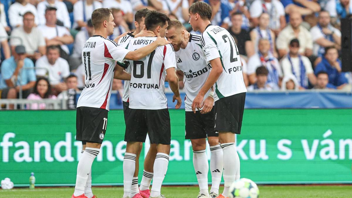 Grupa piłkarzy Legii Warszawa w białych strojach świętuje gola, stojąc blisko siebie i przytulając się.