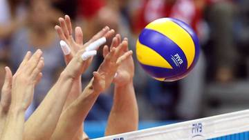 Piłka do siatk&oacute;wki w locie nad siatką, otoczona dłońmi zawodnik&oacute;w.