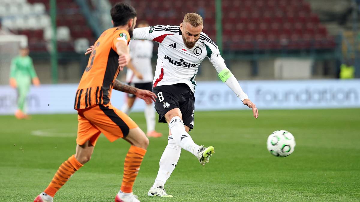 Piłkarz Legii w białej koszulce próbuje kopnąć piłkę, podczas gdy zawodnik drużyny przeciwnej w pomarańczowym stroju stara się mu przeszkodzić.