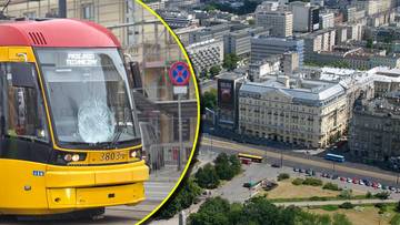 Zbliżenie na przód żółtego tramwaju z widocznymi numerami bocznymi i oznaczeniami. W tle przesunięty obraz panoramy centrum Warszawy z budynkami i ulicami.