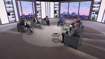 Siedmiu mężczyzn siedzących w kręgu na kanapach w studiu telewizyjnym. W tle okna z widokiem na panoramę miasta nocą.