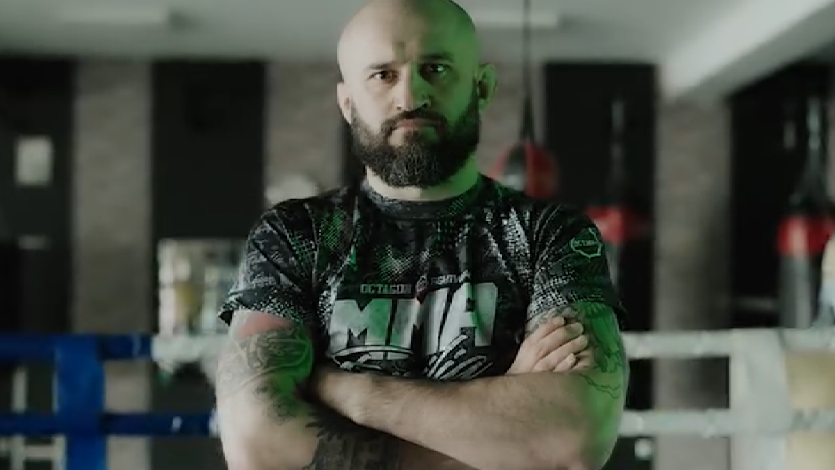 Artur Sowiński, zawodnik MMA, w koszulce z logo MMA, stoi z rękami skrzyżowanymi na piersi w sali treningowej z widocznym ringiem.