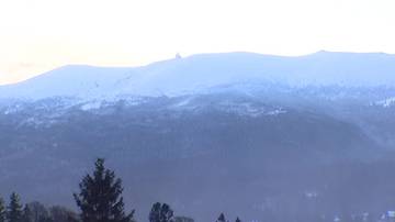 Zimowy krajobraz g&oacute;rski z pokrytymi śniegiem szczytami i widocznym na jednym z nich budynkiem. W tle widać zalesione stoki.