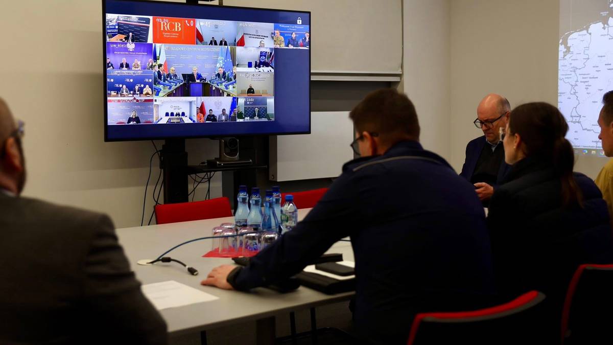 Uczestnicy odprawy hybrydowej patrzą na duży ekran wyświetlający widok wielu os&oacute;b połączonych wideokonferencją.