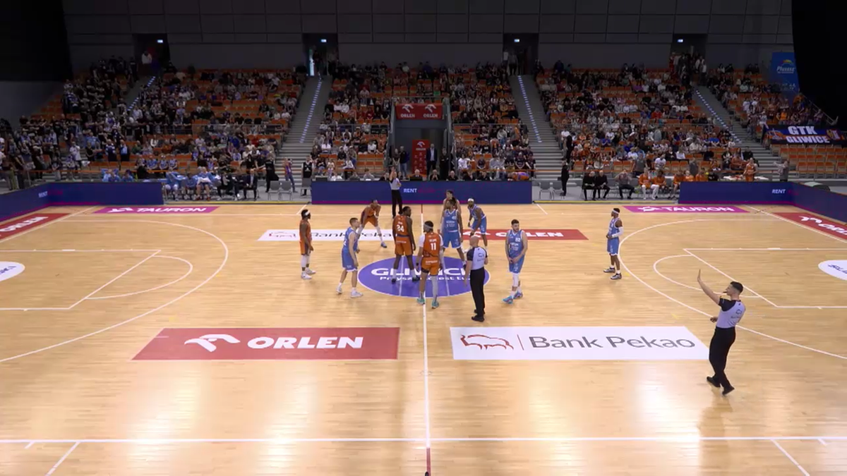 Drużyny koszykarskie Tauron GTK Gliwice i G&oacute;rnik Zamek Książ Wałbrzych ustawione na boisku przed rozpoczęciem meczu, z sędzią wskazującym kierunek gry.