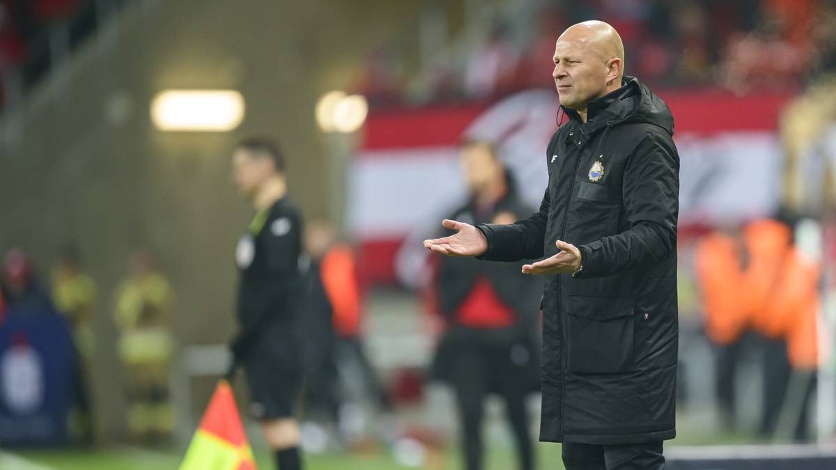 Mężczyzna w czarnym płaszczu zimowym stoi na boisku piłkarskim, gestykulując prawą ręką.