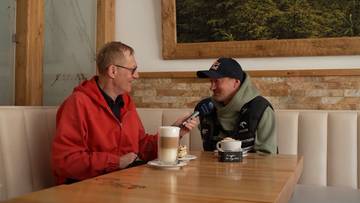 Dw&oacute;ch mężczyzn siedzi przy stoliku w kawiarni. Jeden z nich, ubrany w czerwoną bluzę, trzyma mikrofon i przeprowadza wywiad z drugim mężczyzną, ubranym w czapkę z daszkiem i kamizelkę.