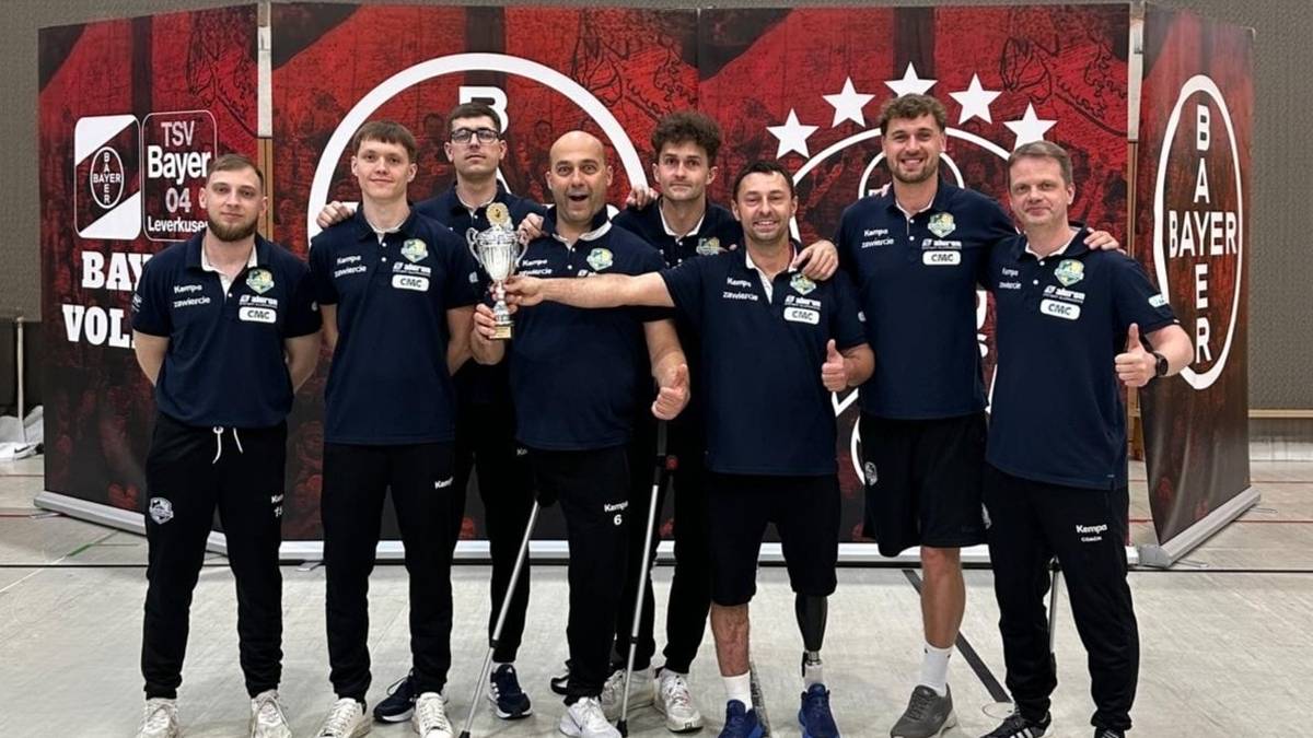 Grupa siedmiu mężczyzn w strojach siatkarskich z niebieskimi koszulkami i czarnymi spodenkami stoi razem, trzymając puchar. Po lewej stronie stoi trener z pucharem w rękach. Wszyscy uśmiechają się.