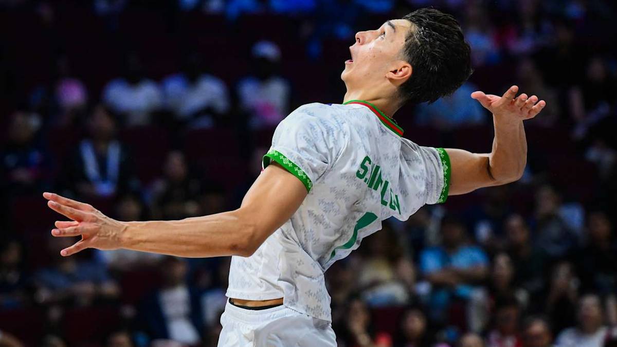 Młody siatkarz w białej koszulce z zielonymi akcentami, z rękami rozłożonymi na boki, wykonuje akcję podczas meczu.