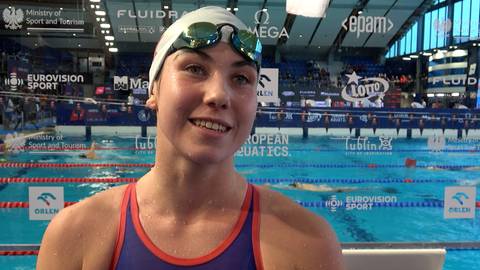 Portret uśmiechniętej młodej kobiety w stroju pływackim, okularach i czepku. W tle basen pływacki z torami i trybunami.