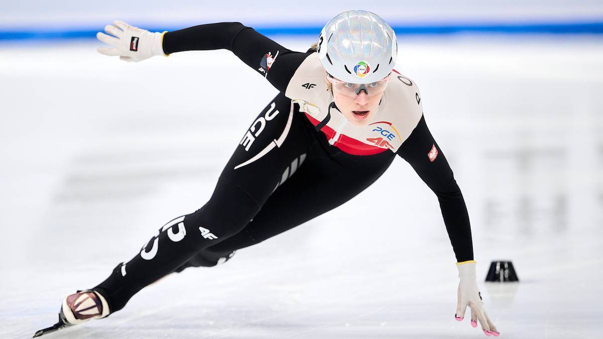 Zawodniczka short tracku w kasku i okularach, ubrana w kombinezon z białymi i czerwonymi elementami, pochyla się do przodu podczas jazdy na lodzie, z rękami wyciągniętymi do przodu i jedną ręką dotykającą lodu.