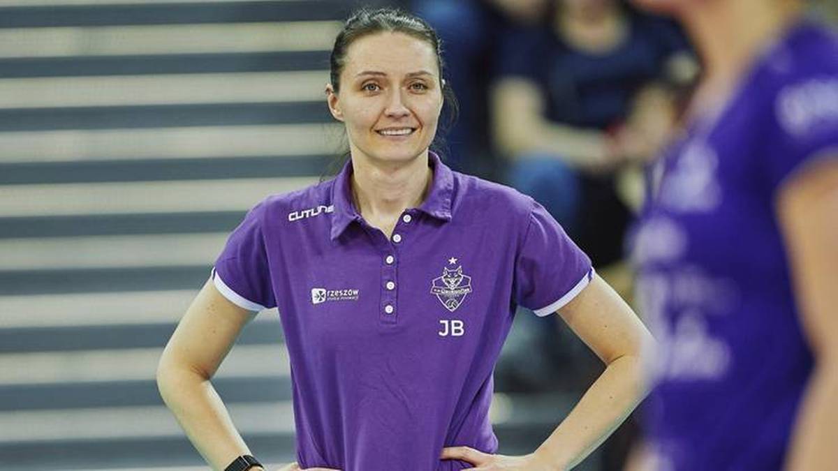 Uśmiechnięta kobieta w fioletowej koszulce polo z rękoma na biodrach, stojąca w hali sportowej.