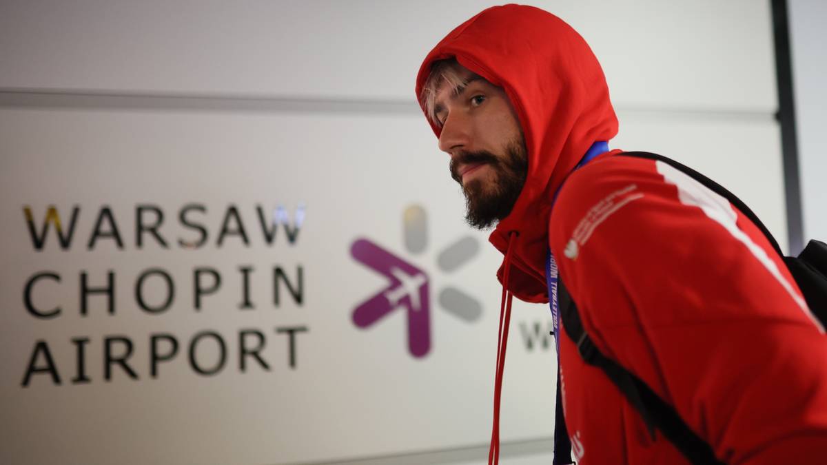 Mężczyzna w czerwonej bluzie z kapturem na tle napisu "WARSAW CHOPIN AIRPORT"