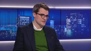 Mężczyzna w okularach i zielonym swetrze na tle niebieskiego ekranu z widokiem na miasto nocą.