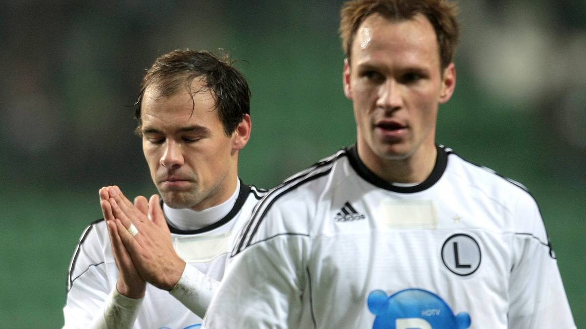 Dw&oacute;ch piłkarzy w białych koszulkach z czarnymi pasami na ramionach, jeden z nich składa ręce jak do modlitwy, drugi patrzy przed siebie.