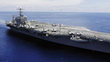 Lotniskowiec USS Abraham Lincoln na oceanie, z widocznymi samolotami na pokładzie i mniejszymi jednostkami pływającymi w oddali.