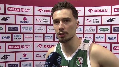 Mężczyzna w zielonej koszulce sportowej z widocznym numerem i herbem klubu na ramieniu, udziela wywiadu. W tle znajduje się ścianka z logotypami sponsor&oacute;w.