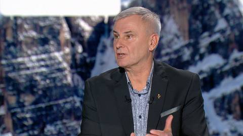 Mężczyzna w średnim wieku z siwymi włosami i w garniturze, m&oacute;wiący do kamery.