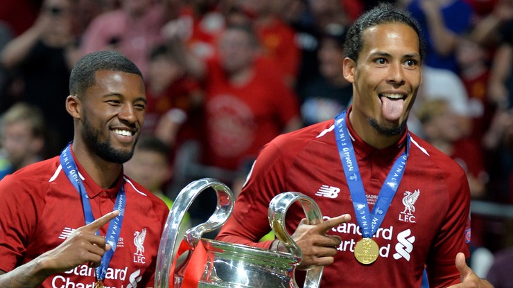Dwóch piłkarzy w czerwonych koszulkach Liverpoolu z medalami na szyi, jeden z nich z trofeum Ligi Mistrzów w dłoniach, pokazuje język.