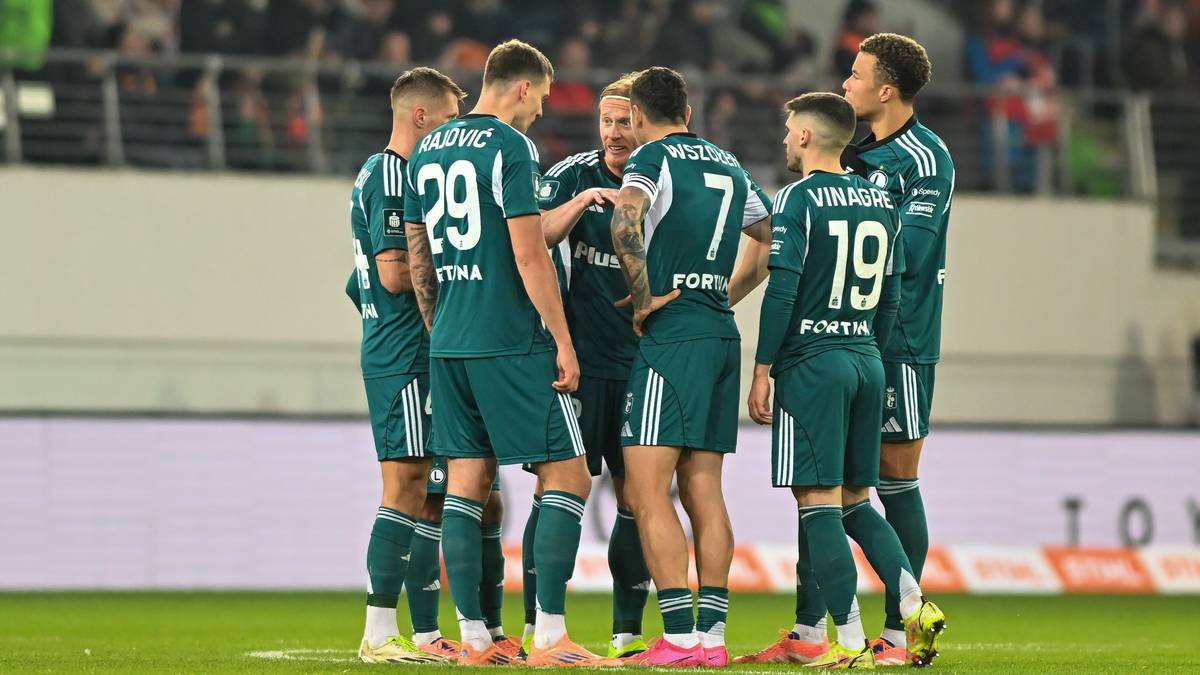 Drużyna piłkarska w zielonych strojach stoi w kółku na boisku.