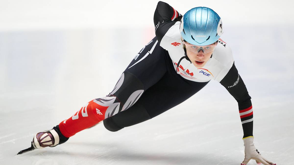 Łyżwiarka szybka w niebieskim kasku i czarno-czerwonym kombinezonie wykonuje zakręt na lodowisku.