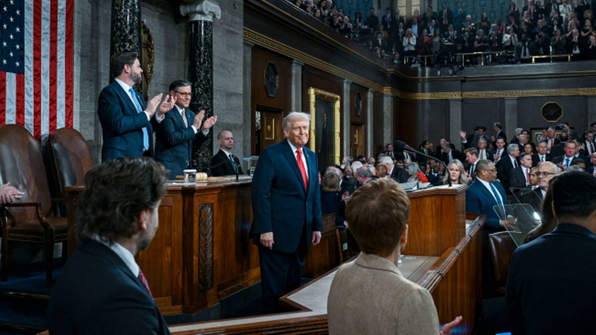 Donald Trump w płaszczu i czerwonym krawacie stoi przed grupą ludzi podczas przem&oacute;wienia w dużej sali z widownią na podwyższeniu.