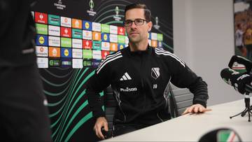 Trener Legii Warszawa siedzi przy stole podczas konferencji prasowej, ubrany w dres z logo klubu.