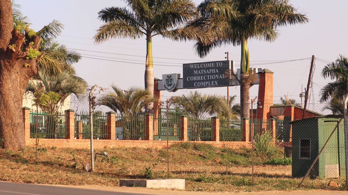 Tablica z napisem "WELCOME TO MANZINI CORRECTIONAL COMPLEX" wisząca nad wejściem do kompleksu, otoczona palmami i budynkami.