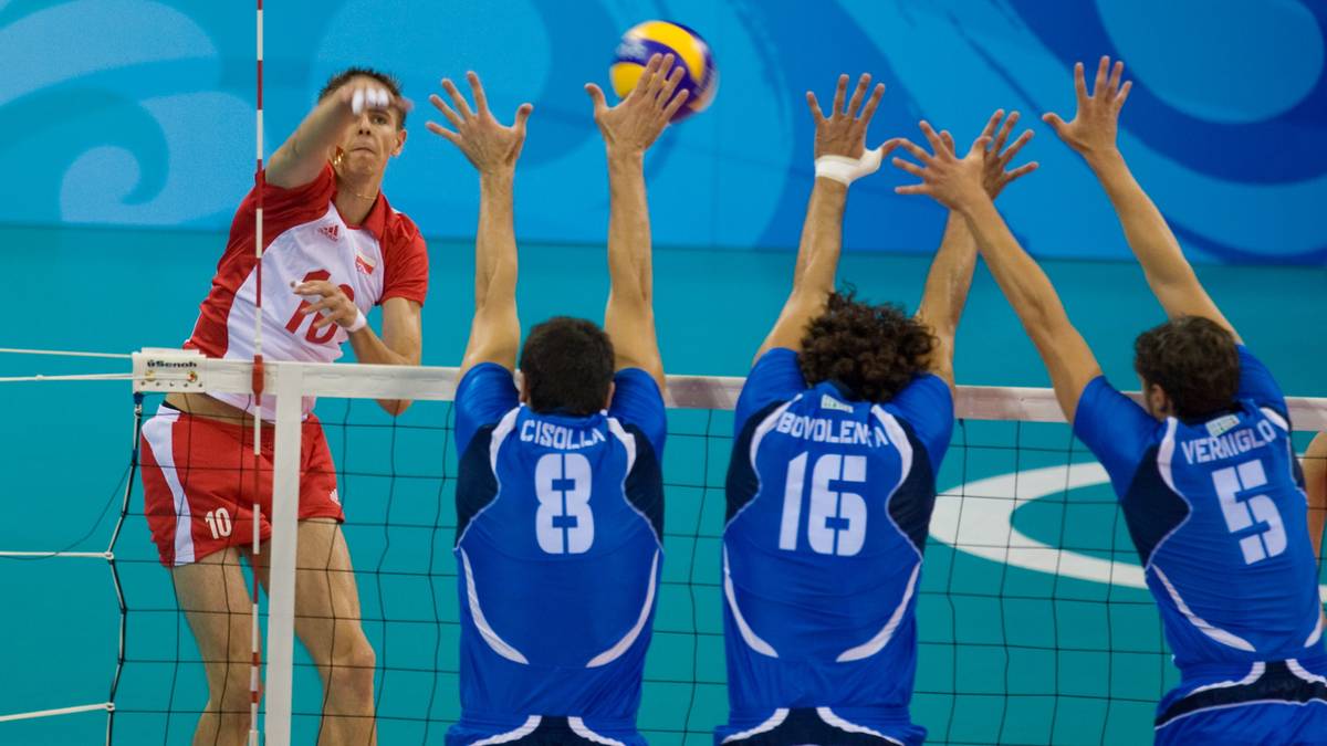 Siatkarz w białej koszulce z czerwonymi rękawami atakuje piłkę, podczas gdy trzech graczy w niebieskich koszulkach próbuje go zablokować.