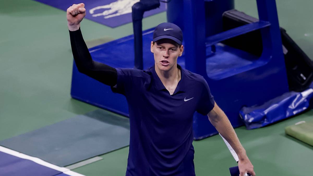 Jannik Sinner, tenisista, w niebieskiej koszulce z krótkim rękawem i czarną czapką z daszkiem, unosi zaciśniętą prawą pięść w geście triumfu na korcie tenisowym.