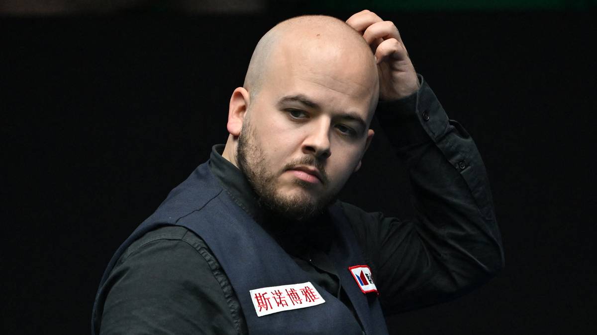 Portret mężczyzny z ogoloną głową i zarostem, w kamizelce snookerzysty, z ręką na głowie.