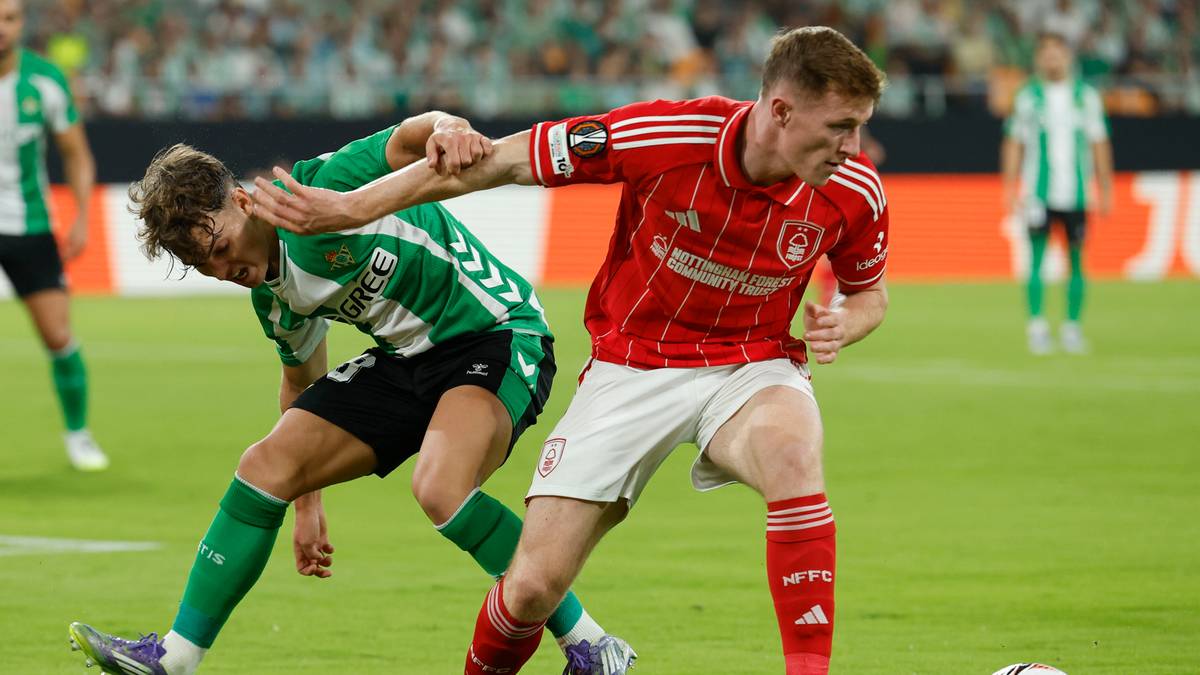 Dwóch piłkarzy w trakcie meczu, jeden w zielono-białej koszulce stara się zatrzymać przeciwnika w czerwono-białej koszulce.