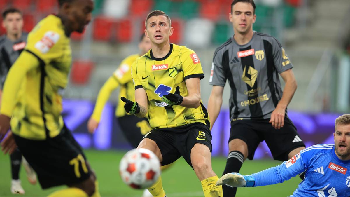 Piłkarze w żółtych strojach walczą o piłkę na boisku, jeden z nich próbuje ją odebrać, podczas gdy bramkarz w niebieskim stroju leży na ziemi.