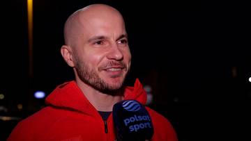 Piotr Michalski, ubrany w czerwoną bluzę, uśmiecha się, trzymając mikrofon Polsat Sport.