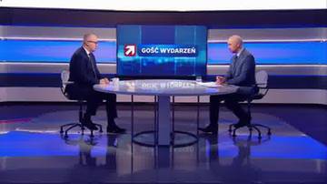 Dwóch mężczyzn w garniturach siedzi naprzeciwko siebie przy okrągłym stole w studiu telewizyjnym. W tle znajduje się duży ekran z logo "GOŚĆ WYDARZEŃ".