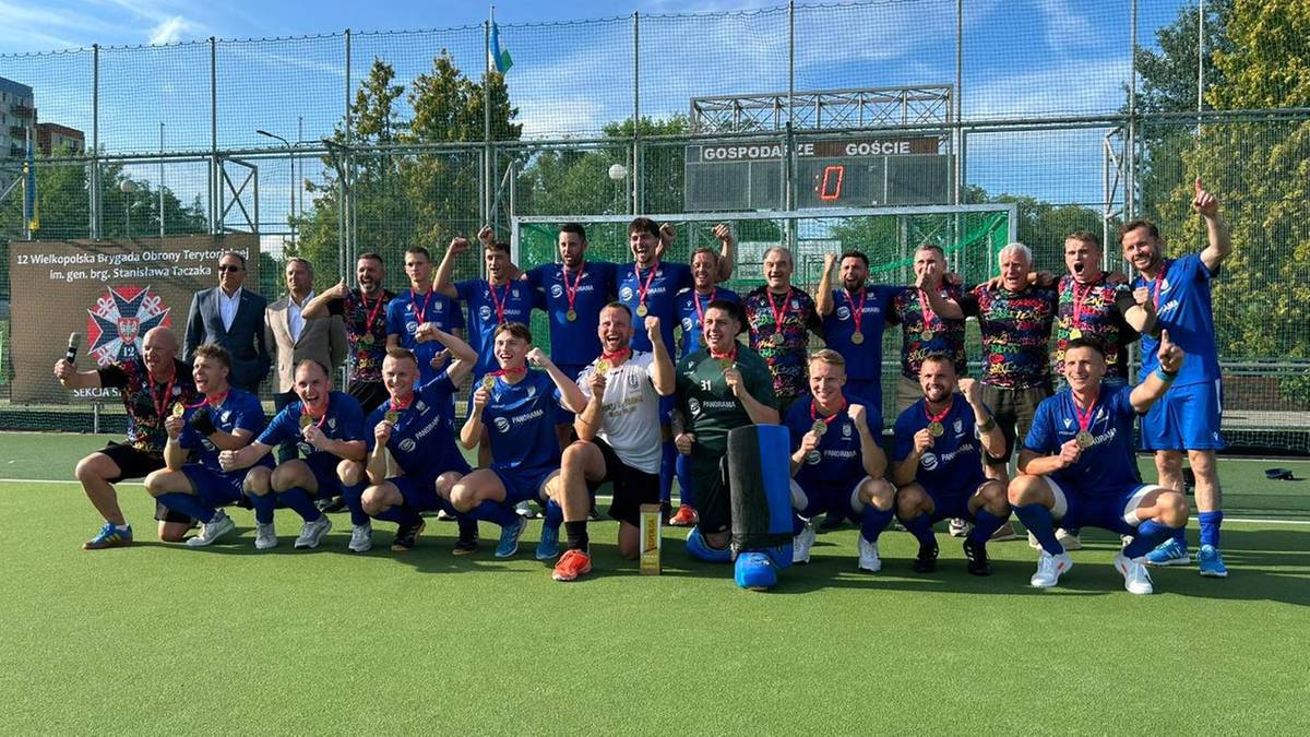 Drużyna hokeja na trawie pozuje do zdjęcia po meczu, wielu zawodników świętuje i pokazuje radość, niektórzy trzymają medale.