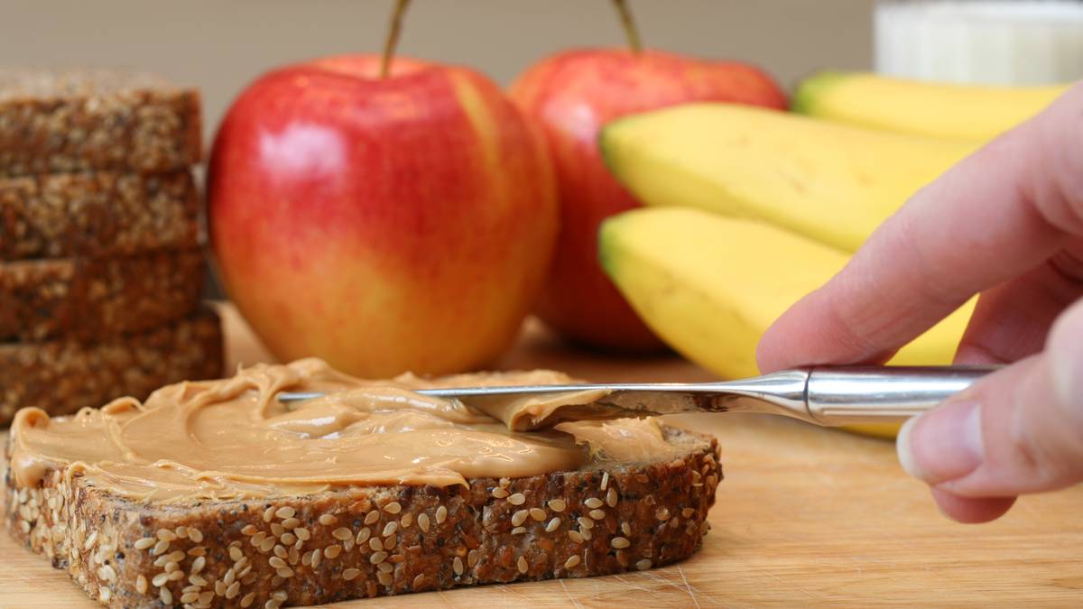 Ktoś rozsmarowuje masło orzechowe na kromce ciemnego chleba razowego. W tle widać jabłka i banany.