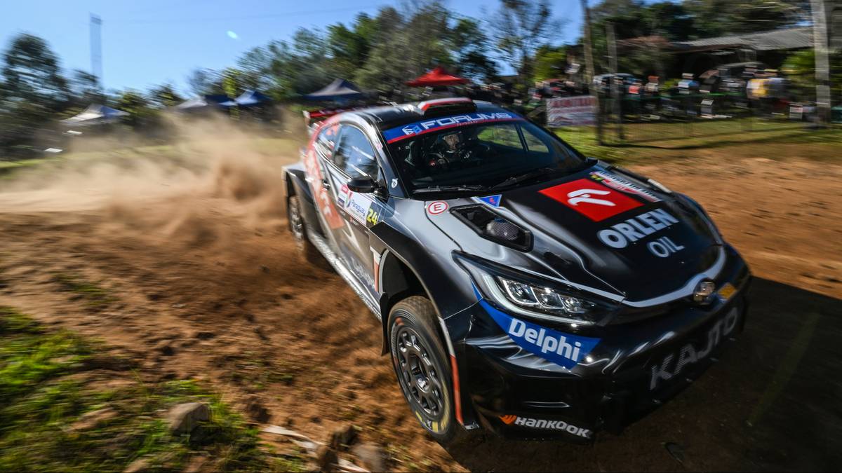 Samochód rajdowy z widocznym logo ORLEN OIL, jadący po piaszczystej drodze i wzbijający tumany kurzu.