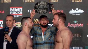 Dwóch zawodników MMA stoi naprzeciw siebie podczas ważenia, a pomiędzy nimi arbiter trzyma pas mistrzowski Babilon MMA nad ich głowami.
