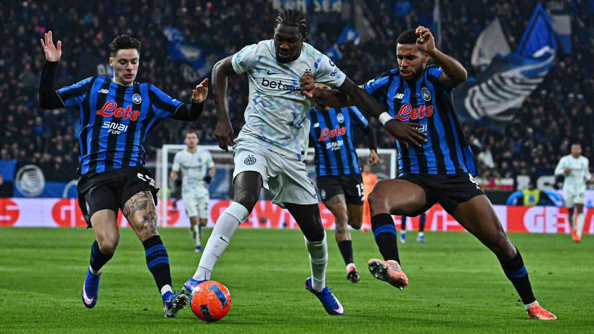 Trzech piłkarzy na murawie stadionu podczas meczu piłki nożnej, dw&oacute;ch w niebiesko-czarnych strojach i jeden w jasnym, walczący o piłkę.