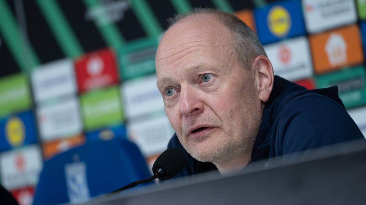 Portret mężczyzny w średnim wieku, m&oacute;wiącego do mikrofonu, z kamiennym wyrazem twarzy.