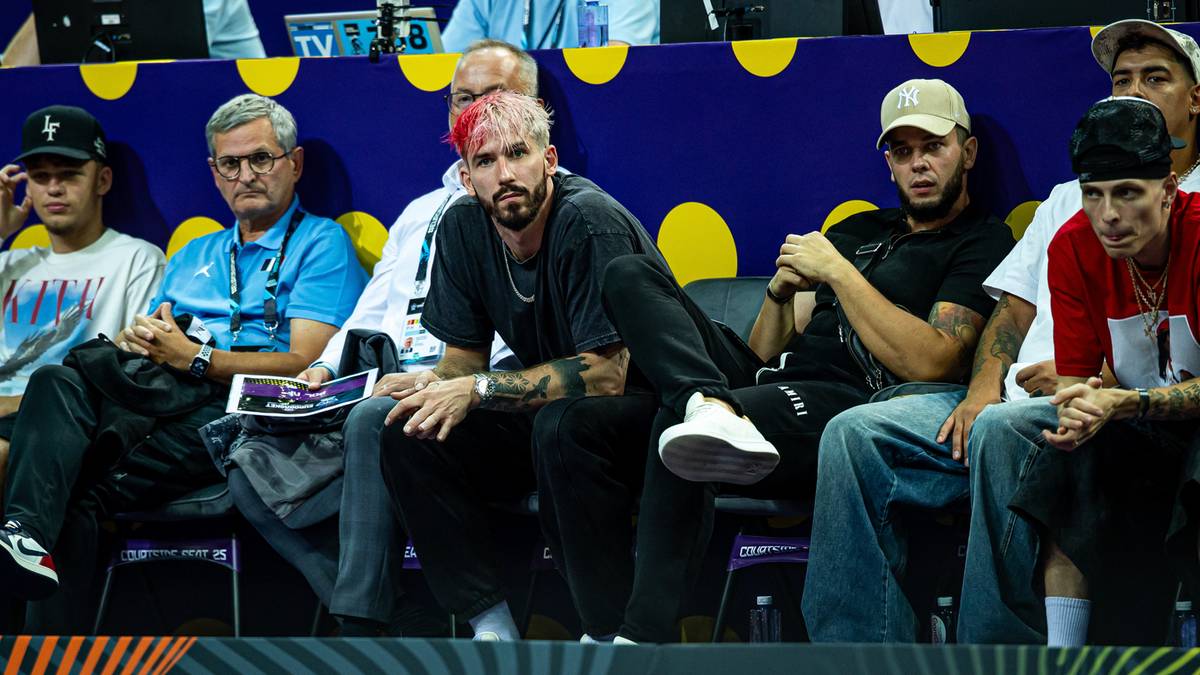 Mężczyzna z r&oacute;żowymi włosami i brodą siedzi na trybunach obok innych os&oacute;b.