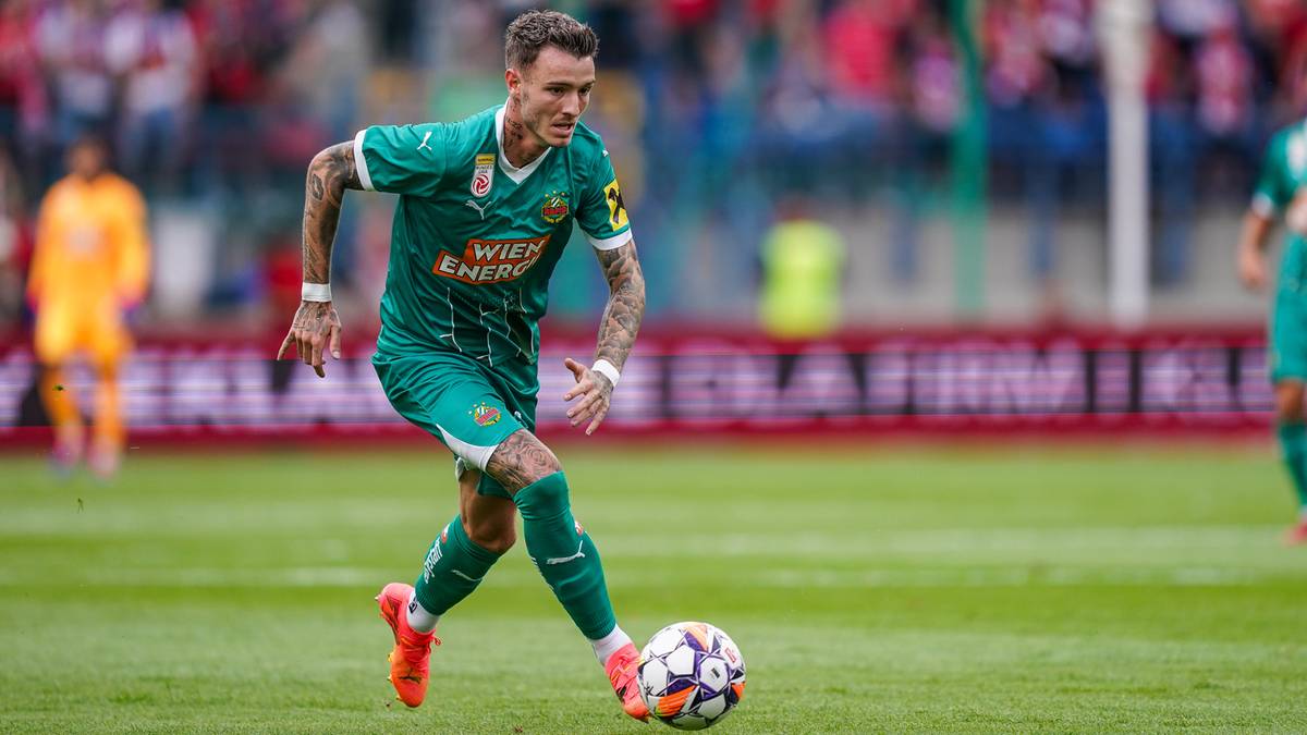 Piłkarz w zielonym stroju z pomarańczowymi elementami biegnie z piłką po boisku, skupiony na grze.