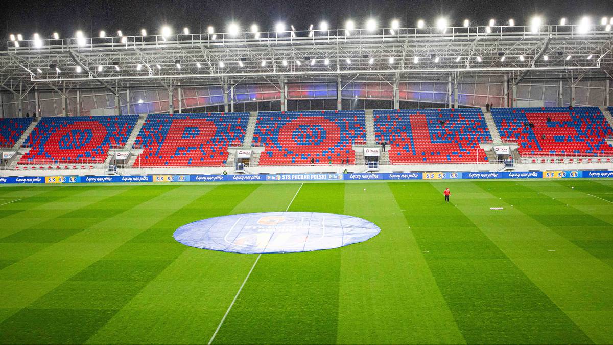Stadion piłkarski z trybunami w kolorach czerwonym i niebieskim, na murawie znajduje się plandeka.
