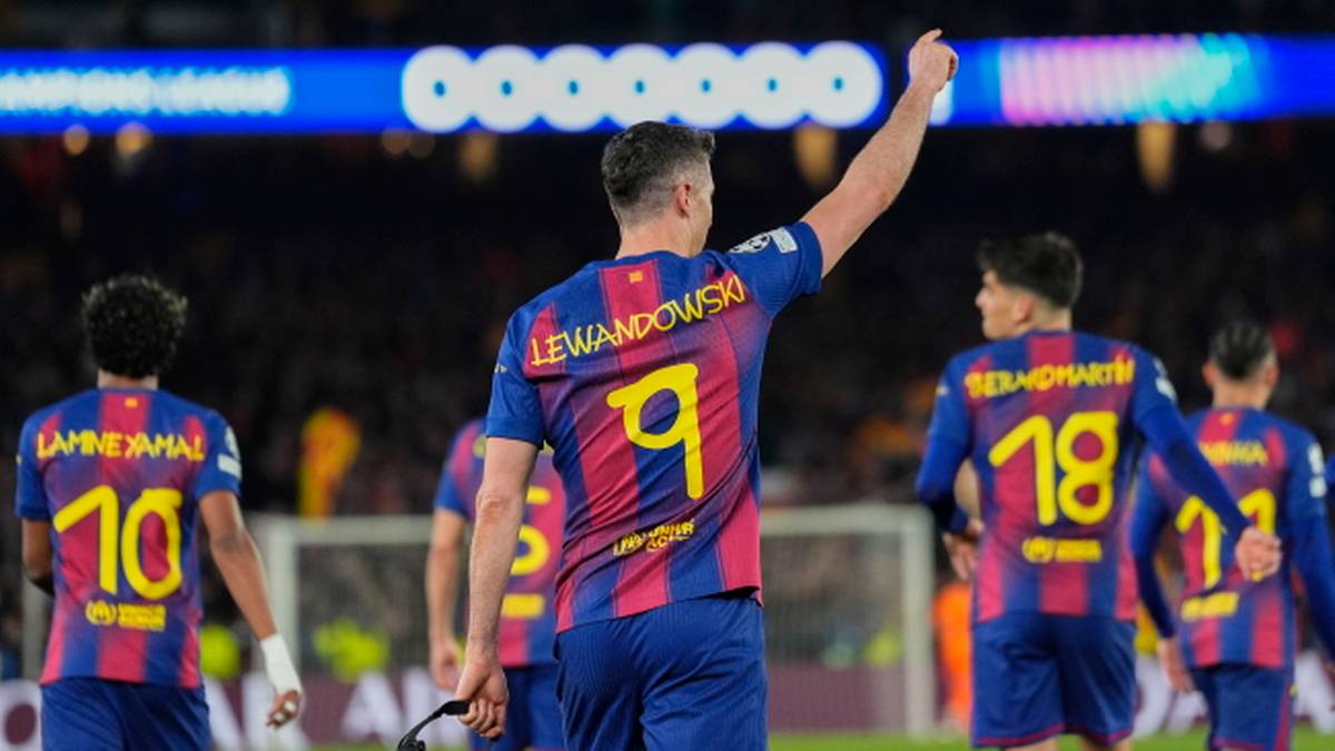 Piłkarz Robert Lewandowski w koszulce FC Barcelony z numerem 9 na plecach, z uniesioną prawą ręką, na tle innych zawodnik&oacute;w drużyny.