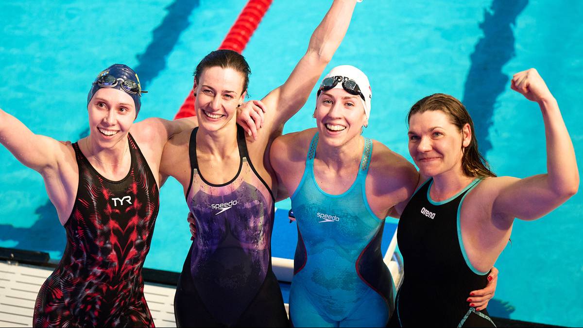 Cztery uśmiechnięte kobiety w strojach pływackich pozują do zdjęcia na brzegu basenu.