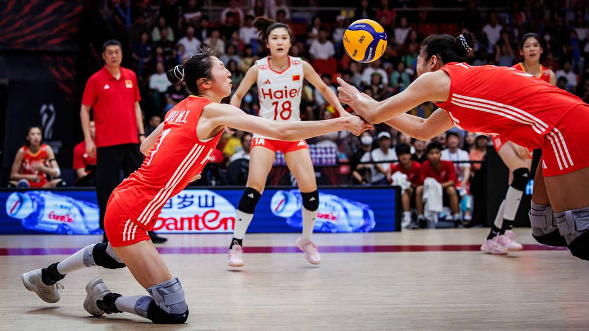 Dwie zawodniczki siatkówki próbują odbić piłkę w locie; jedna z nich klęczy na parkiecie, a druga wyciąga ręce.