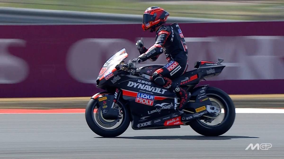 Motocyklista w czerwono-czarnym kombinezonie i kasku jedzie po torze wyścigowym obok tablicy wynik&oacute;w z napisem "FINISH".