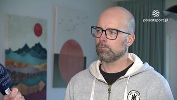 Mężczyzna z brodą i okularami w szarym swetrze z kapturem.